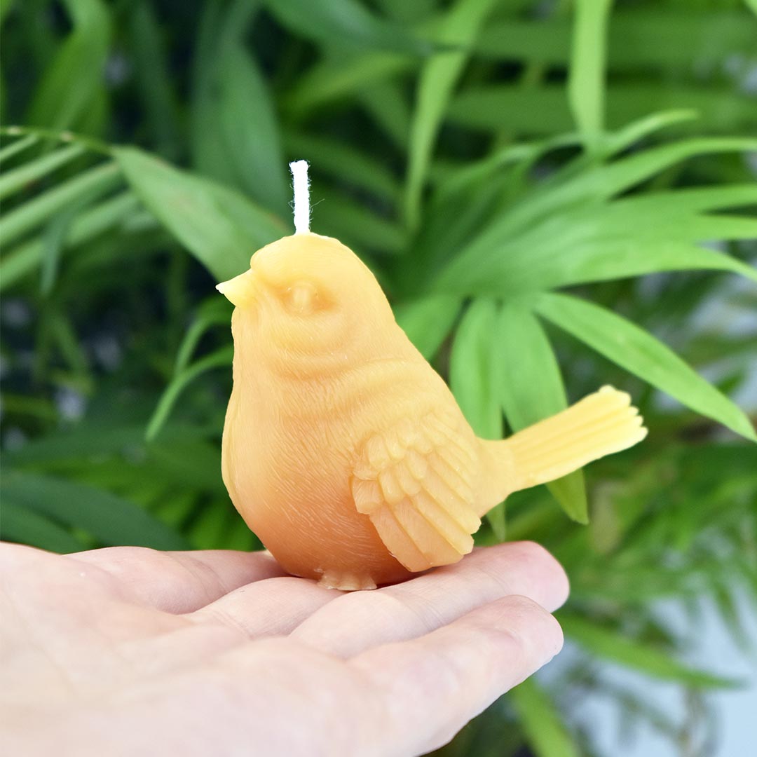 A small sparrow shaped beeswax candle perched on a hand with green foliage in the background. Cute little bird candle.